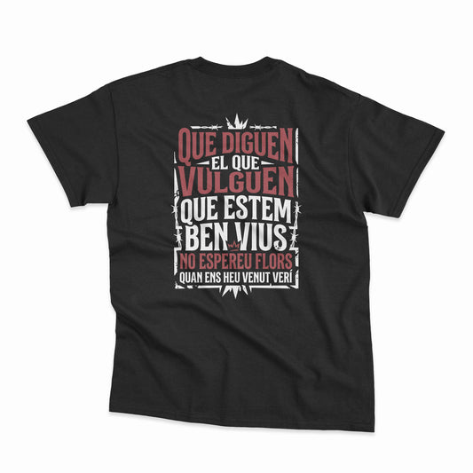 Samarreta unisex negra amb la frase "Que diguen el que vulguen, que estem ben vius, no espereu flors quan ens heu venut verí" de La Gossa Sorda a l'esquena, de Canya de Sucre.