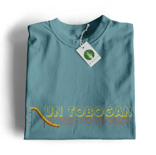 Samarreta infantil color blau dusty amb la frase “Li falta un tobogan” i il·lustració d’un tobogan, disseny original de Canya de Sucre.